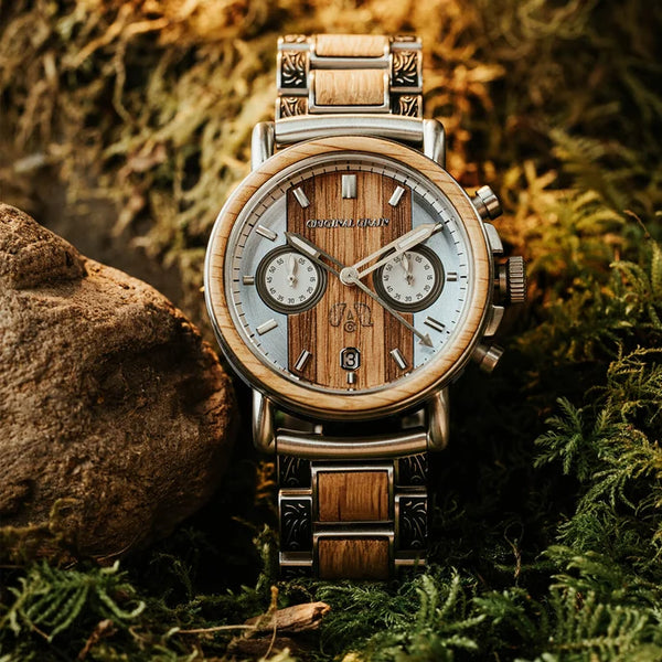 A wristwatch with a wood-grain dial and matching wood-and-metal link bracelet, photographed outdoors among moss, ferns, and a rock. The watch face has two small subdials and a silver-toned case.