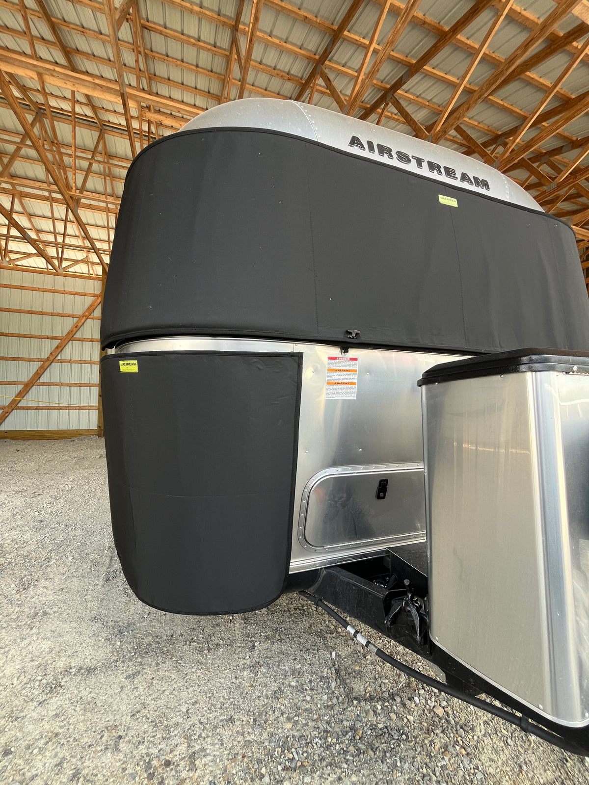 A silver Airstream trailer with black Airstream Travel Trailer Padded Rock Guard Covers over the front windows is parked inside a wooden, metal-roofed storage building on gravel.