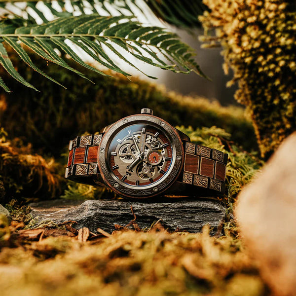 A luxury skeleton wristwatch with a dark wood and metal link bracelet, resting on a dark stone surrounded by moss, ferns, and natural forest elements.
