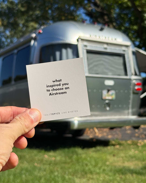 A hand holds a square Airstream TableTopics Game card asking about the inspiration behind choosing the trailer, held in front of a parked silver camper.