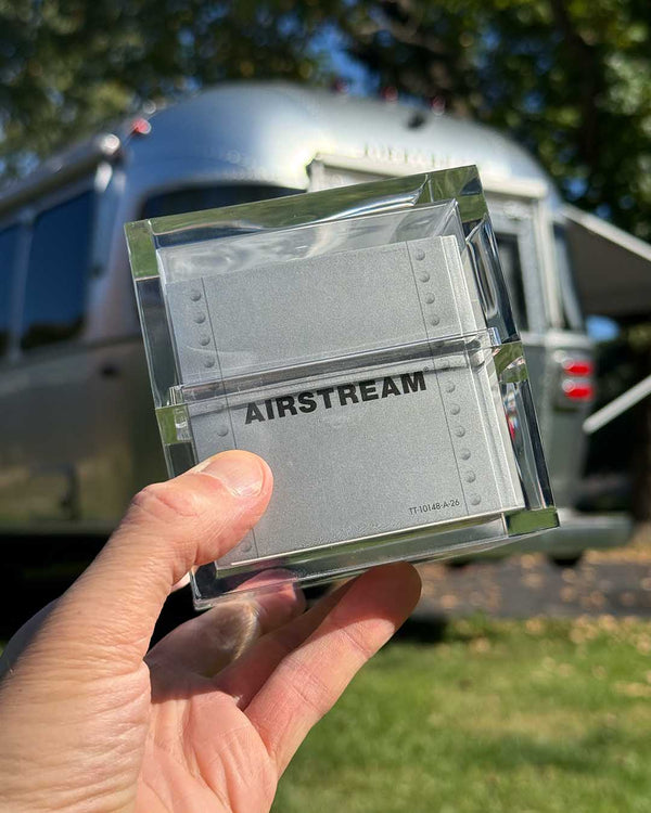 A hand holds a square, clear acrylic case containing an Airstream TableTopics Game with metallic, rivet-patterned cards in front of a silver travel trailer.