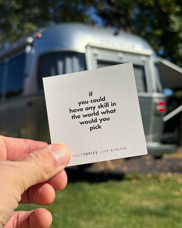 A hand holds an Airstream TableTopics Game card that reads, If you could have any skill in the world what would you pick, in front of a parked travel trailer.