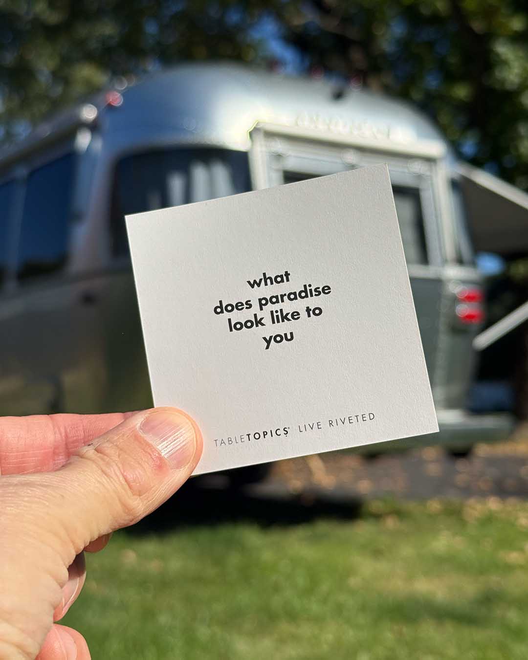 A hand holds a square Airstream TableTopics Game card that reads, what does paradise look like to you, with a silver Airstream trailer blurred in the background.