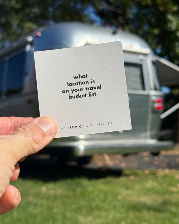 A hand holds a square Airstream TableTopics Game card asking for a bucket list location, with a silver trailer blurred in the background.