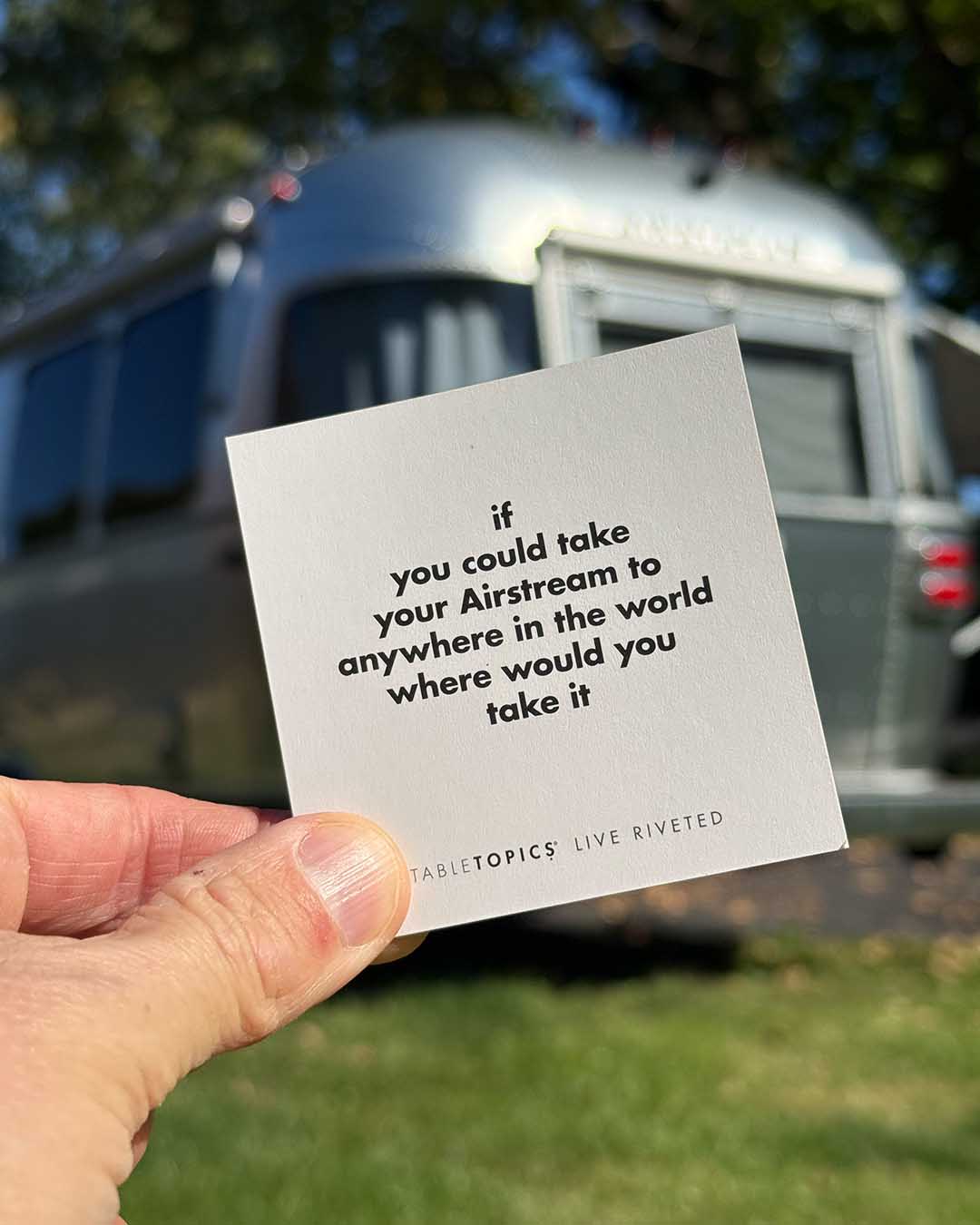 A hand holds a square Airstream TableTopics Game card that reads, If you could take your Airstream to anywhere in the world where would you take it, with a travel trailer blurred in the background.