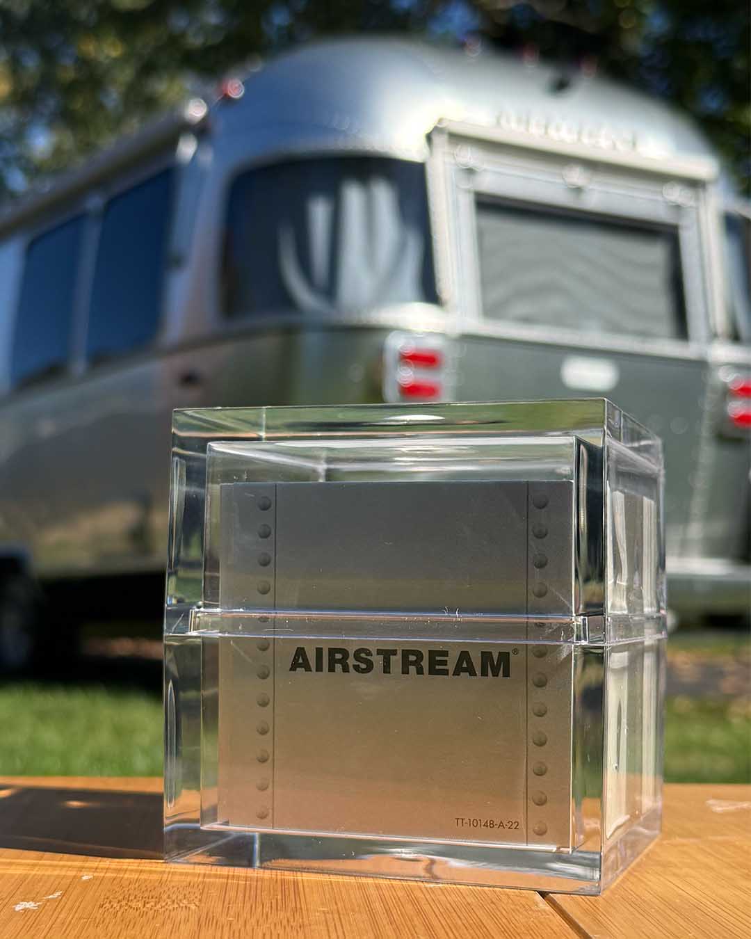 A square, clear acrylic box containing the Airstream TableTopics Game with metallic cards, sitting on a wooden surface with a travel trailer in the background.