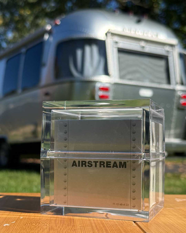 A square, clear acrylic box containing the Airstream TableTopics Game with metallic cards, sitting on a wooden surface with a travel trailer in the background.