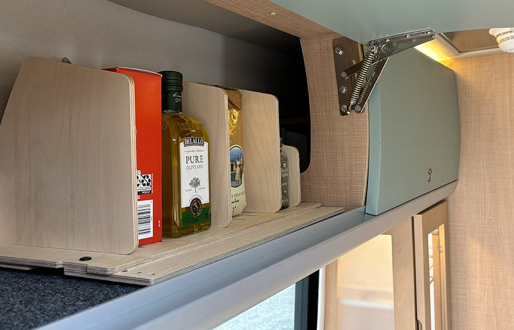 An Airstream Touring Coach Roof Locker Organization unit by Airworthy Equipment Company stores olive oil, a red box, and additional pantry items in the kitchen area. Positioned beside a blue overhead cabinet featuring a visible hinge mechanism that exemplifies efficient overhead storage, the scene includes light brown walls and a window in the background.