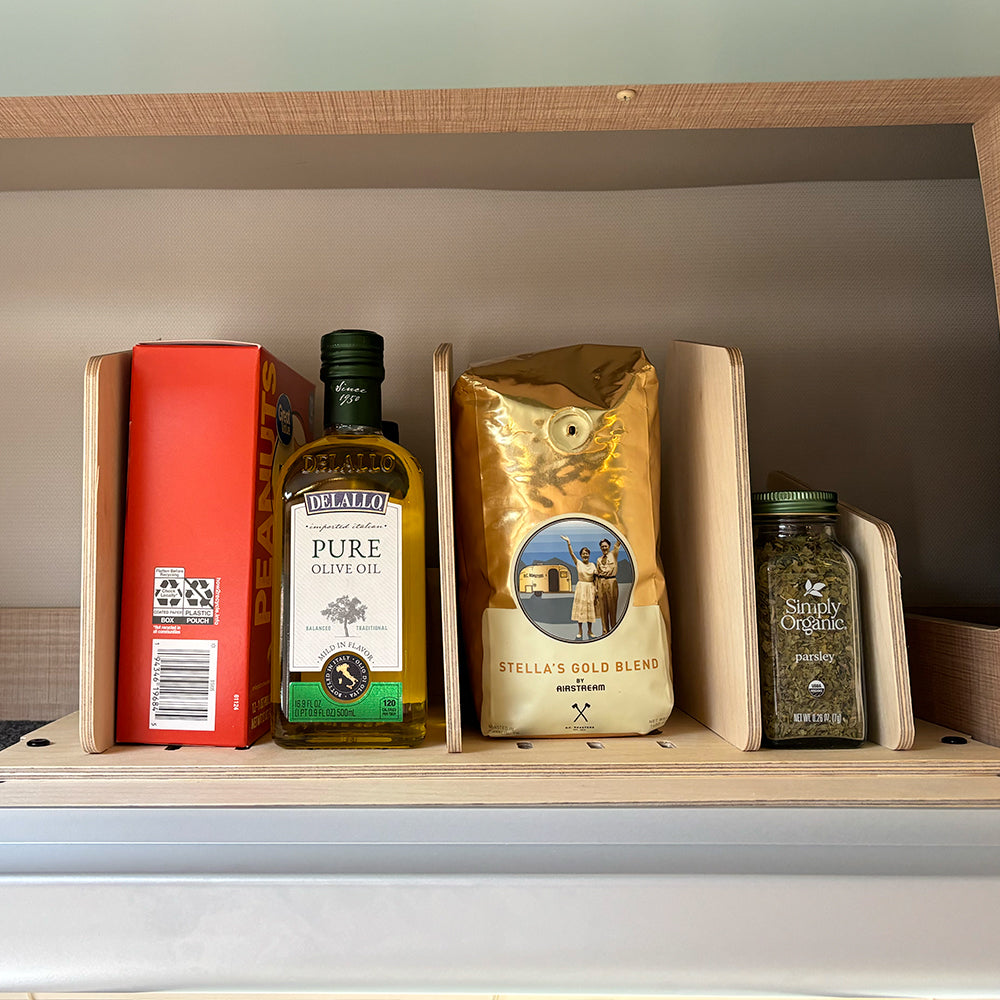 Four pantry essentials—a red box of peanuts, a bottle of olive oil, a gold bag of coffee, and a green jar of dried herbs—are neatly separated by wooden dividers on the shelf, creating an ideal organization system with the Airstream Touring Coach Roof Locker Organization by Airworthy Equipment Company for maximizing space in your roof locker.