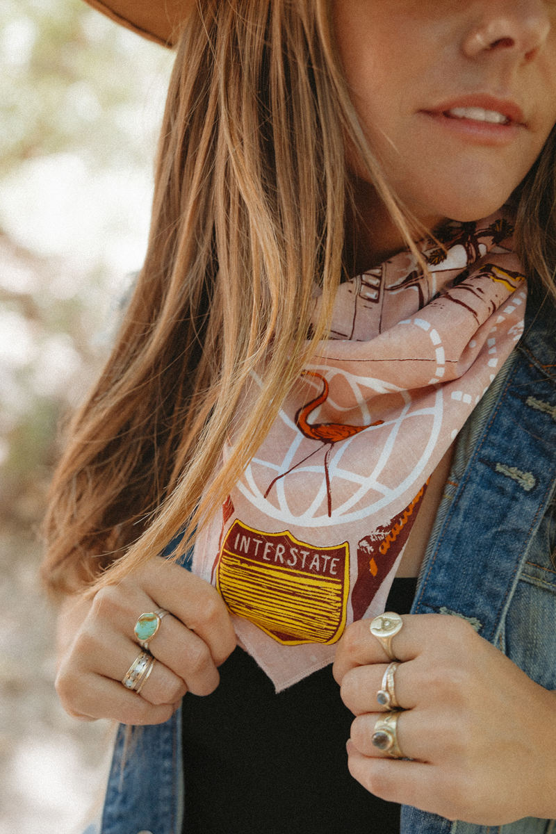 A woman wearing rings and a denim jacket adjusts a scarf with an Interstate sign design. Her face is partially visible, and she has long hair and a hat.