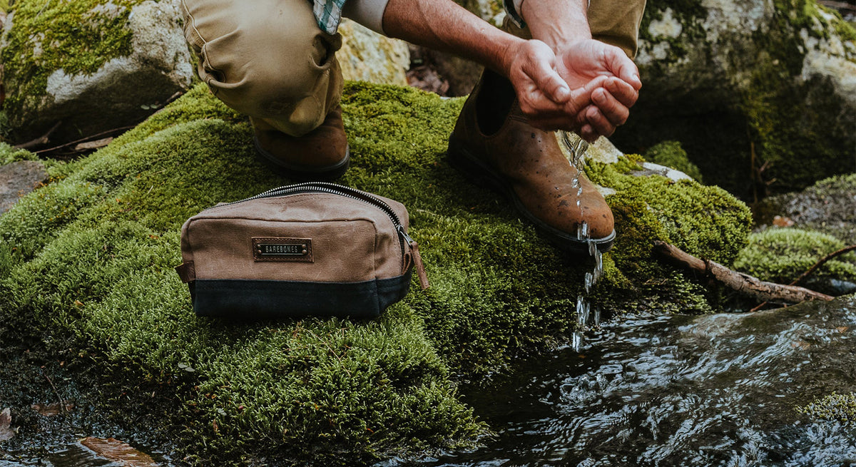 A person kneels on mossy rocks by a stream, scooping water with their hands. A brown zippered pouch rests nearby.