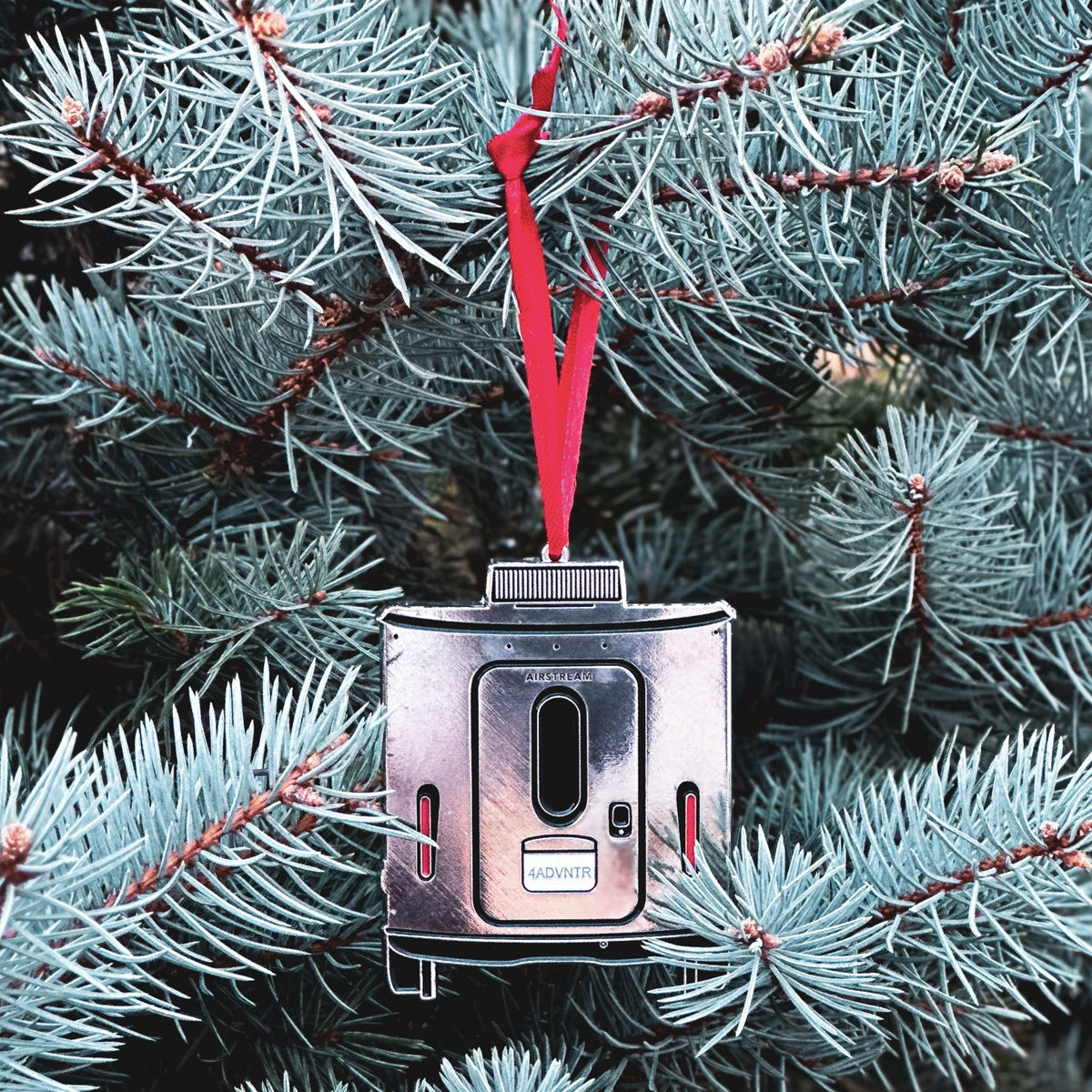 A shiny Airstream Basecamp Trailer Flat Ornament with a red ribbon hangs from the branch of a pine tree, nestled among green needles—perfect for travel lovers and adventurers at heart, evoking the excitement of hitting the open road.