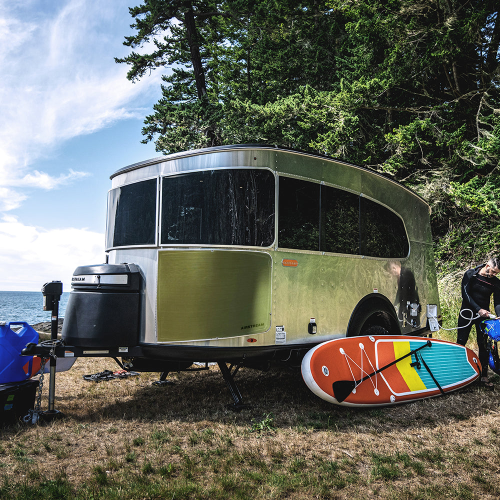 A silver Airstream camper trailer, featuring the minimalist Rock Guard Rock Wraps Logo Collection for Basecamp X, is parked on grass near the water with trees in the background. A colorful paddleboard and gear are arranged on the ground in front of the trailer.