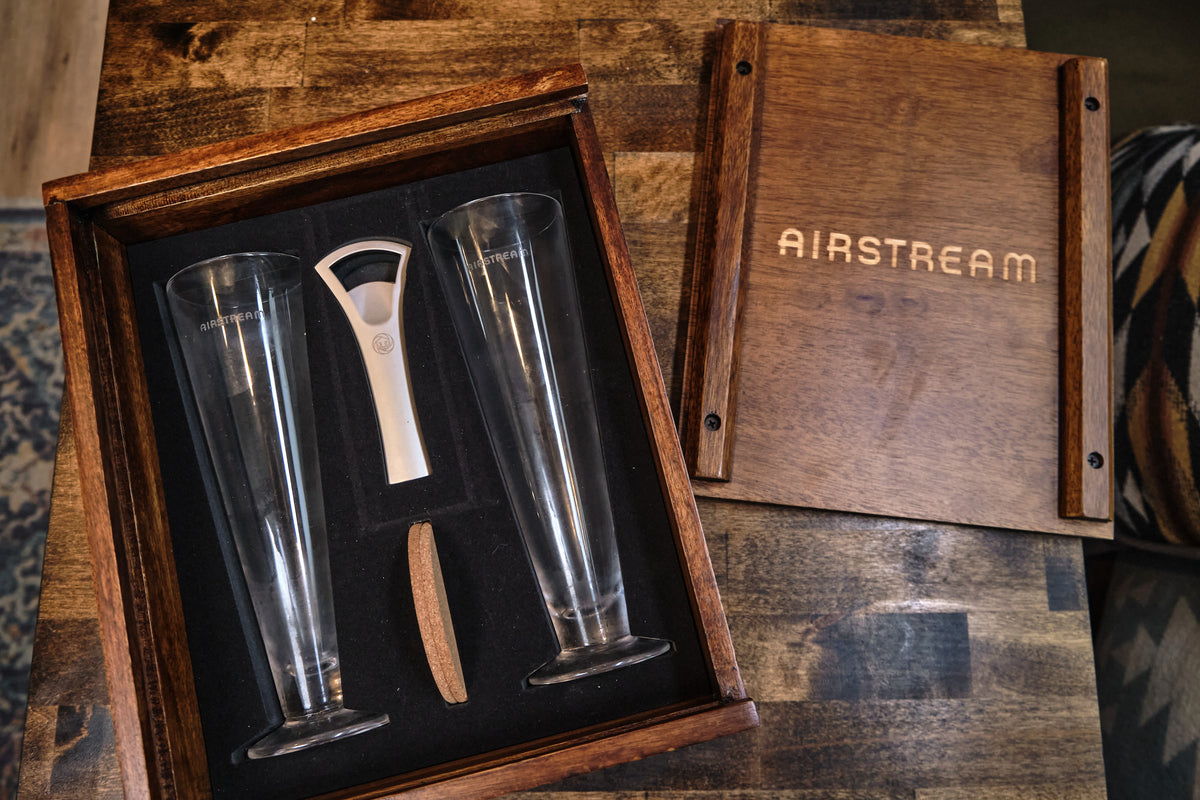 The Airstream Beer Glass Gift Set is elegantly displayed in an Acacia wood case labeled AIRSTREAM, featuring two tall beer glasses, a deluxe bottle opener, and a cork—all neatly arranged on a wooden surface.