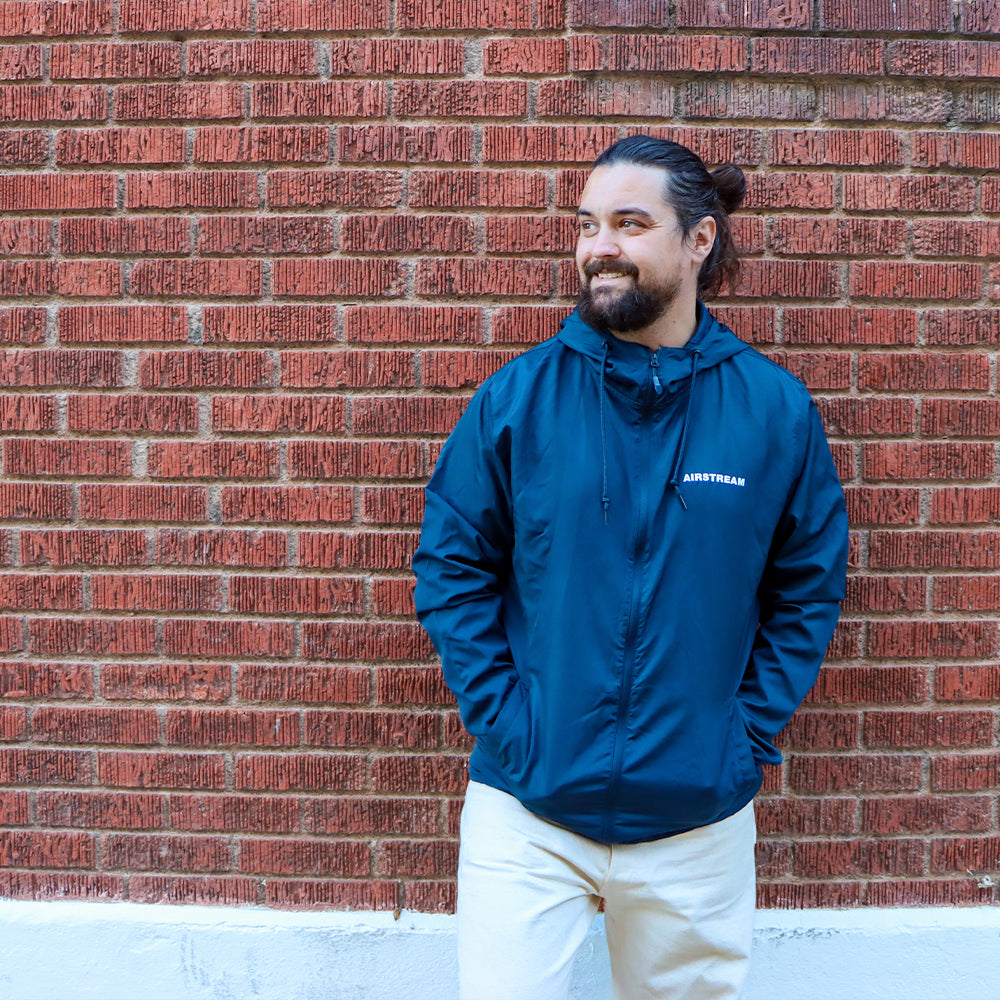 A man with a beard and long hair in a bun, wearing the Airstream Windbreaker Full-Zip Jacket in navy and light pants, stands in front of a red brick wall, smiling and looking to the side.
