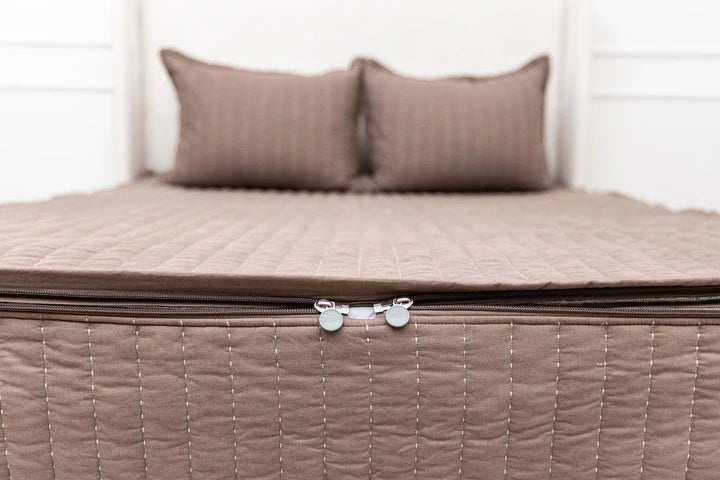A neatly made bed featuring the Airstream RV Fit Beddy’s for Trade Wind Travel Trailers—a brown quilted zippered bedding set with a convenient zipper at the foot—and two matching pillows, all set against a white headboard.