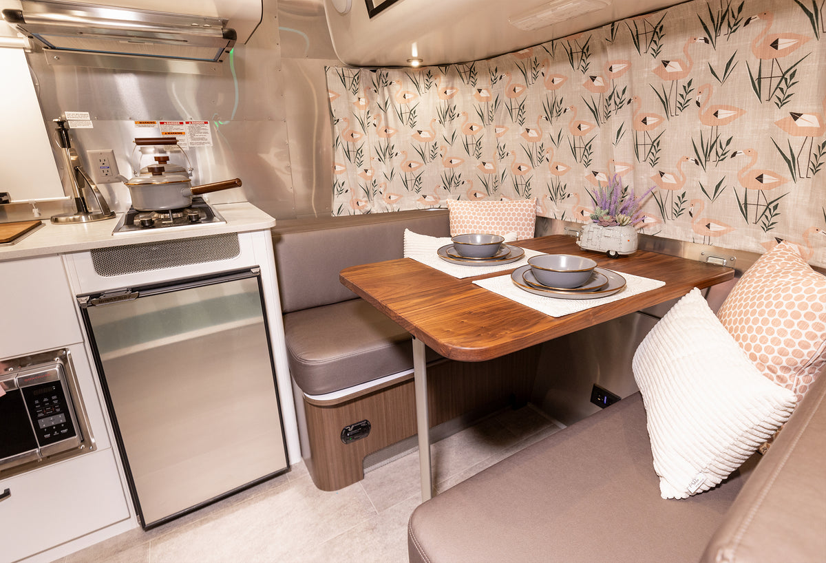 Modern RV kitchen and dining area featuring a premium Airstream Wood Dinette Table Upgrade for Flying Cloud Travel Trailers set for two, complemented by brown cushions, a small fridge, stove, microwave, and floral curtains. Neutral tones create a cozy and inviting space.