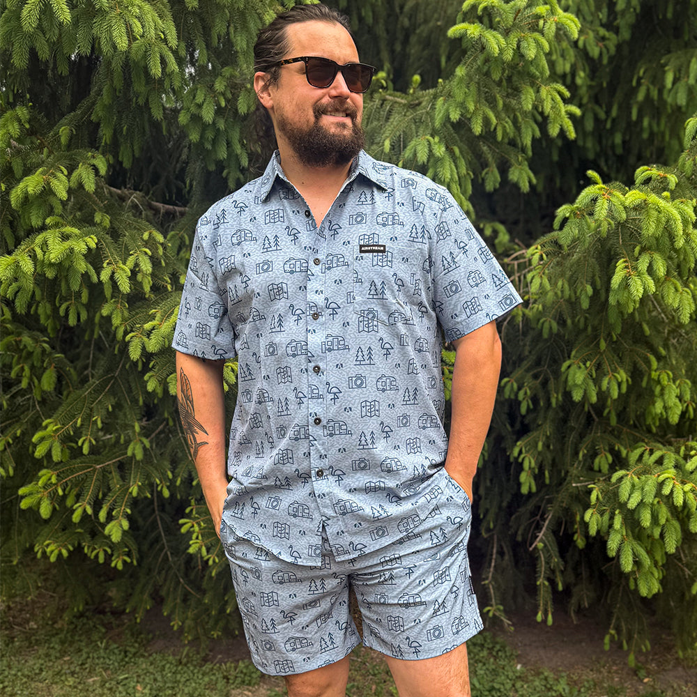 A man wearing sunglasses and a matching blue patterned shirt and light blue Airstream Conversational Shorts stands outdoors in front of green pine trees, smiling with his hands in his pockets.