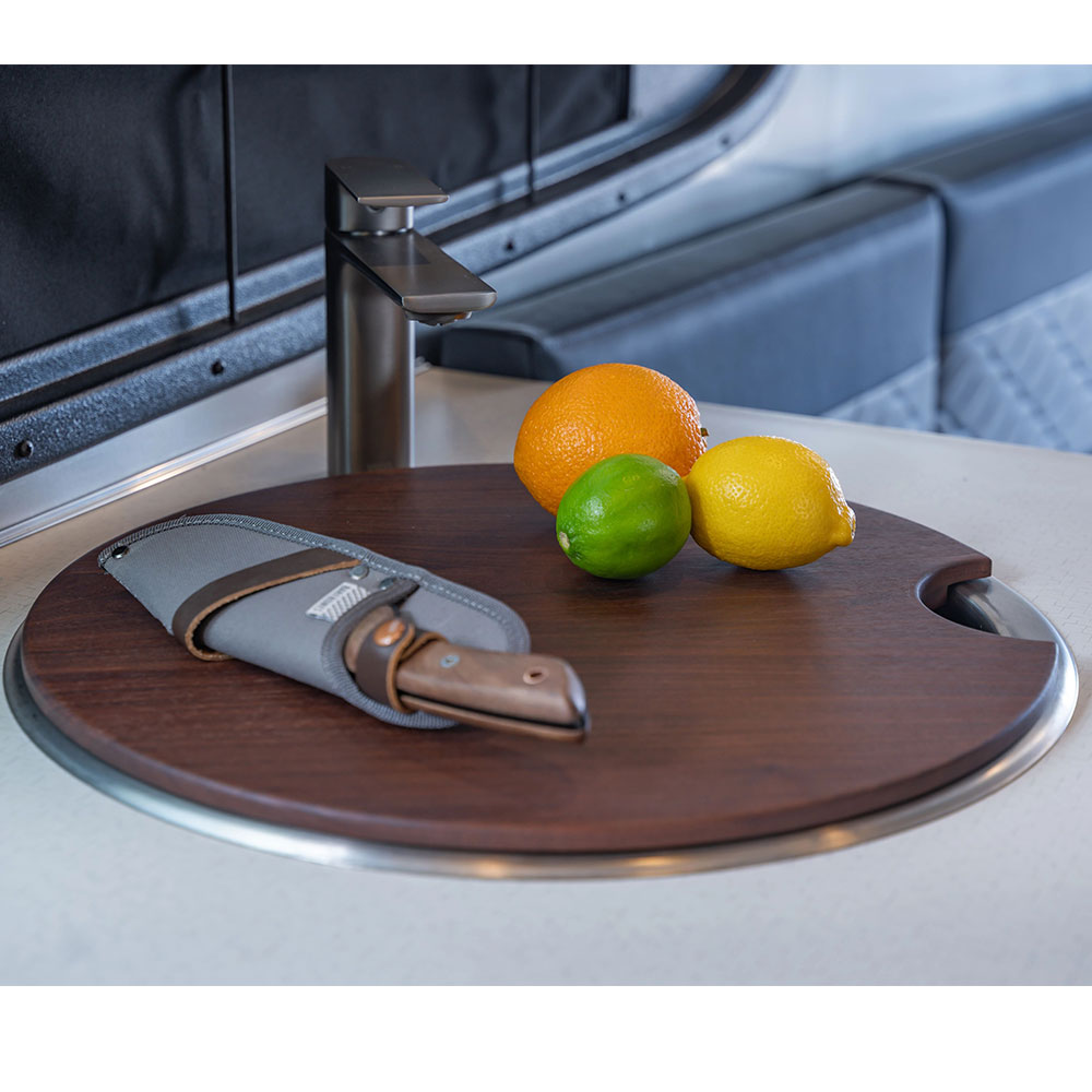 An Airstream Custom Sink Cutting Board for Basecamp, complete with a knife in a sheath and fresh citrus fruits—an orange, a lime, and a lemon—rests on a white countertop next to a modern faucet, making it the perfect accessory for your Airstream Basecamp kitchen.