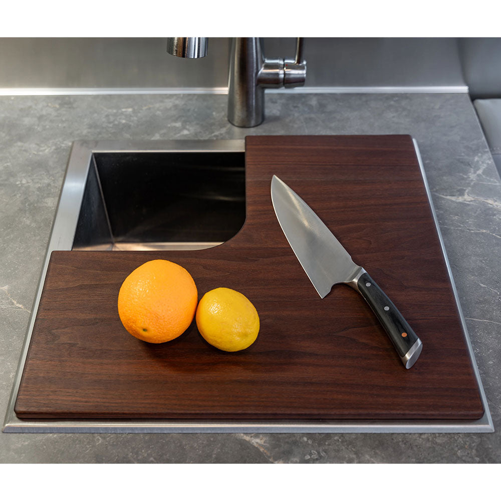 An Airstream Custom Sink Cutting Board for International Travel Trailers covers part of the kitchen sink, with an orange, a lemon, and a chef’s knife resting on top. The countertop beneath is gray with a marble-like pattern.