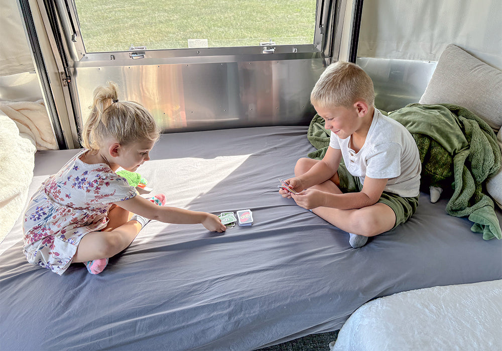 Two young children sit cross-legged on a bed inside a Victorinox travel trailer, playing a card game together. Sunlight pours through the window, illuminating the cozy space adorned with soft pillows and Airstream fitted sheets. Nearby, blankets and Airstream Dinette Fitted Sheets for Victorinox Travel Trailers add extra comfort.