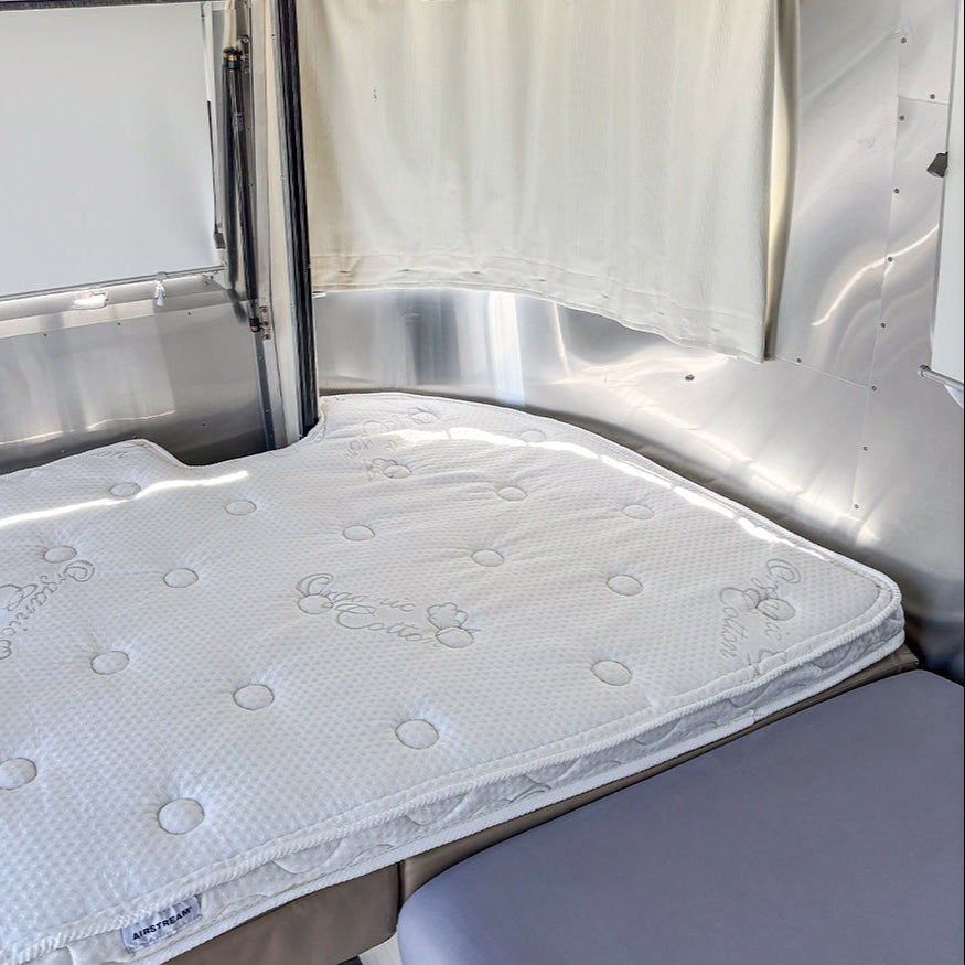 A uniquely shaped white Airstream Dinette Memory Foam Topper, designed for Tommy Bahama Travel Trailers and featuring a textured surface, is placed inside a metallic Airstream Travel Trailer next to a gray seat and a window covered by a light curtain.