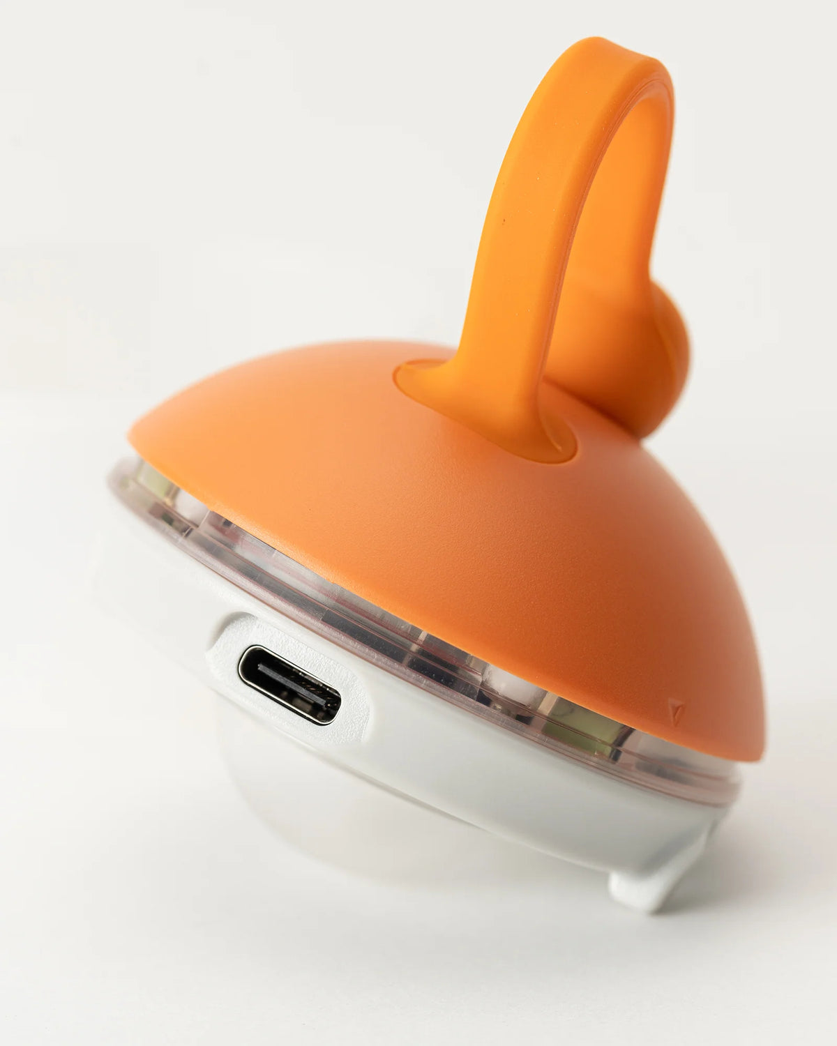 A close-up of the Snow Peak Rechargeable Mini Hozuki, an orange and white lantern with a handle and a visible USB-C charging port on its white base, designed for convenient campsite lighting.