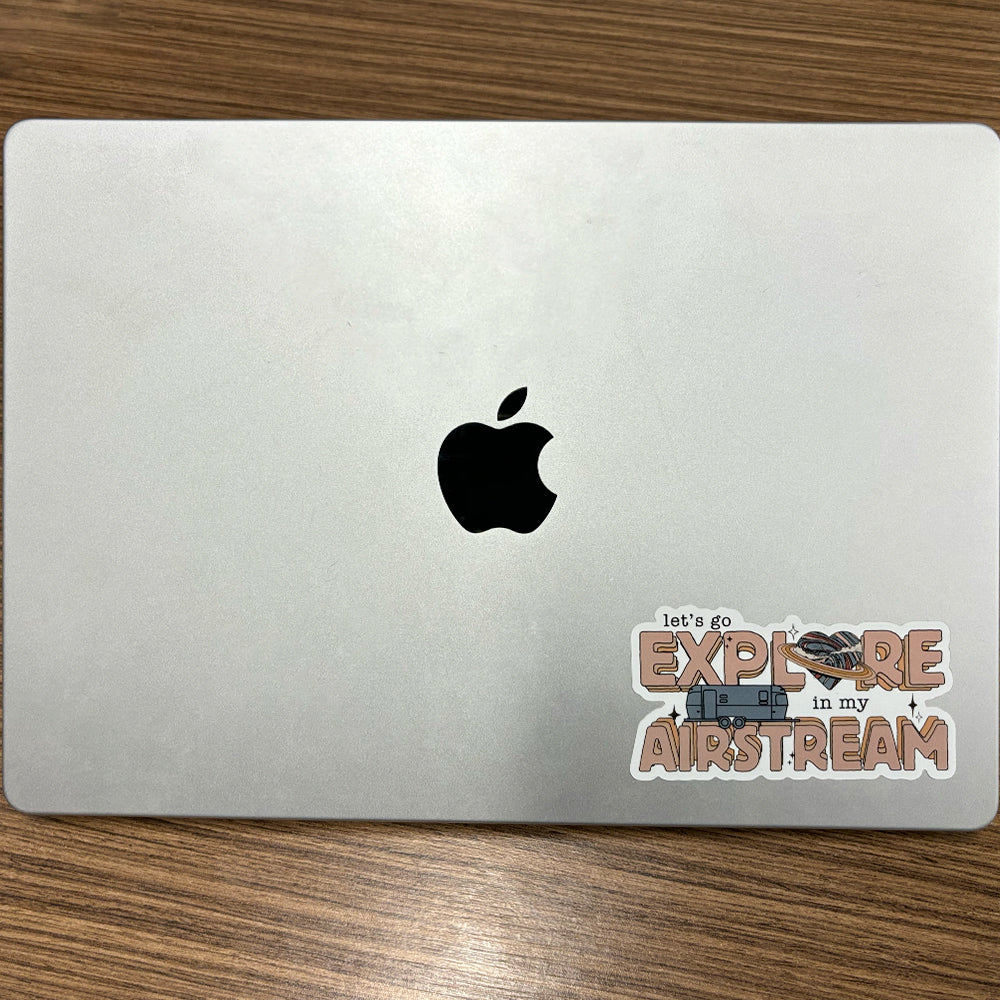 A closed silver MacBook with an Apple logo in the center is adorned in the lower right corner with the Airstream Airstream Let’s Go Explore Sticker, which features an Airstream trailer, nature graphics, and the phrase “lets go explore in my Airstream”—perfect for anyone who enjoys adding a touch of adventure to their gear.