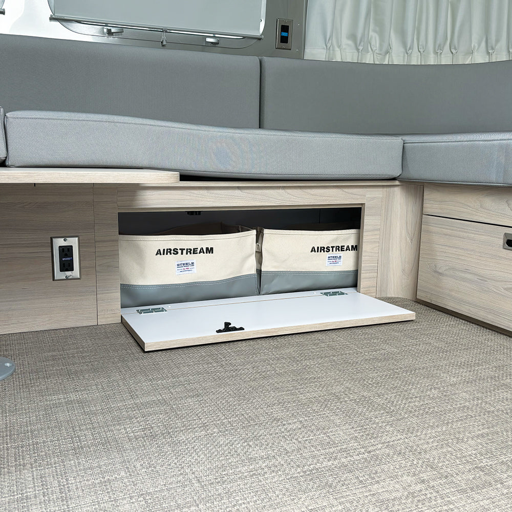 An open storage compartment beneath the L-shaped cushioned bench in a modern Globetrotter 30′ RV reveals two Airstream x Steele Canvas 30′ Globetrotter Lounge Storage Bins, set against light wood paneling and beige carpet flooring.