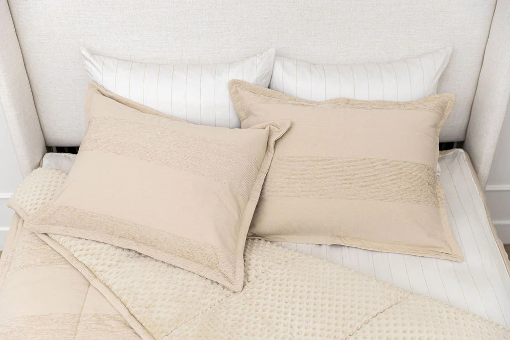 Two beige decorative pillows rest on a bed adorned with the Airstream RV Fit Beddy’s for Excella Travel Trailers, paired with white striped pillows and a textured beige blanket to create a cozy and neutral RV bedding arrangement.