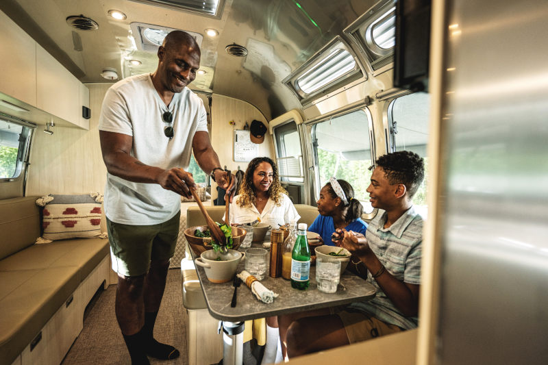 A smiling family of four enjoys a meal together inside a modern RV. The father serves salad while the mother and two children sit at a dining table, laughing and talking. Sunlight streams in through the windows.