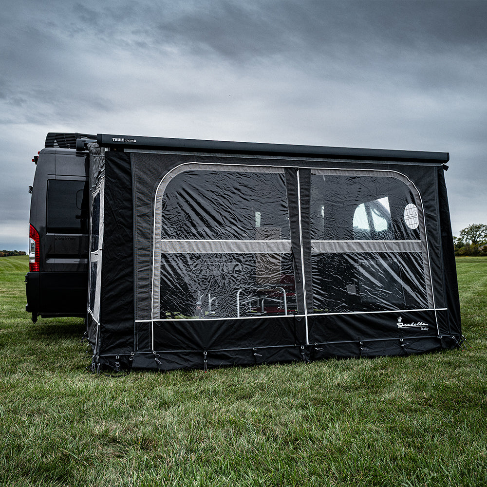 A black van with an attached Buddy Awning Enclosure for Airstream Rangeline stands on a grassy field under a cloudy sky. The enclosure features clear panoramic windows, revealing chairs and a table inside.