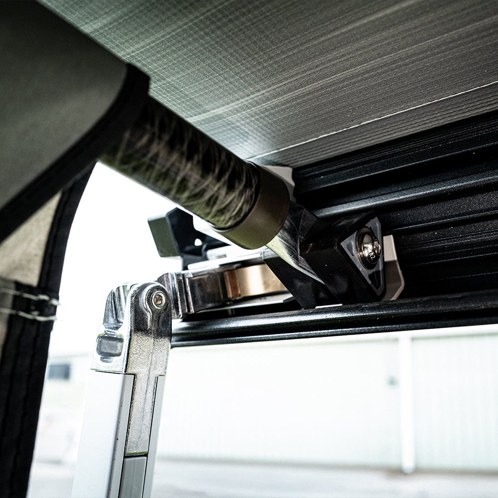 Close-up of the metal hinge and mounting bracket used to secure a rod to the black frame of the Buddy Awning Enclosure for Airstream Rangeline, with panoramic windows and a blurred industrial building visible in the background.