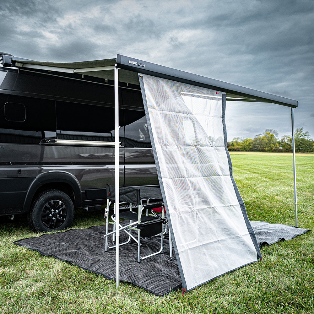 The Shade Screen for Airstream Rangeline is set up on a black van parked on a grassy field, creating excellent sun protection over two folding chairs and a table arranged on a mat under a cloudy sky—perfect for outdoor relaxation.