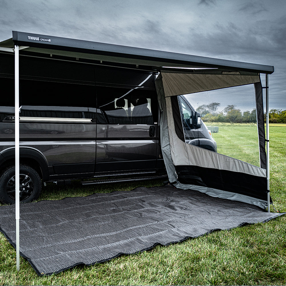 A black camper van with an extended awning and side panel is parked on the grass. The ground beneath the awning is covered by the Outdoor Mat by Isabella, offering a clean and comfortable area to relax, while cloudy skies can be seen in the background.