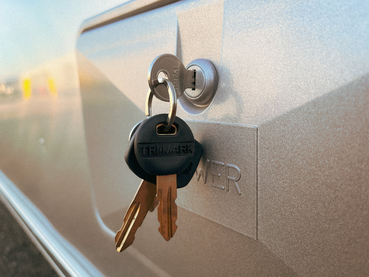 Close-up of two Airstream Water Fill Pre-Cut CH751 Keys inserted in a lock on a metallic surface, with the word “POWER” engraved nearby. The keys, suitable as Airstream replacement keys, are attached to a black plastic keychain labeled “TRIMARK.”.