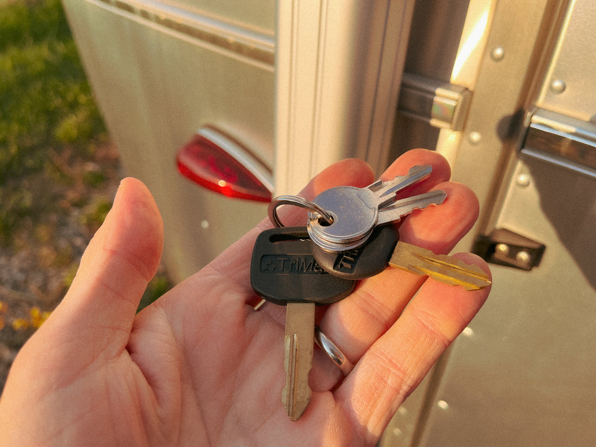 A hand holds several Airstream Pre-Cut Keys for Main Door on a keyring near a metallic door, with sunlight casting warm tones and a blurry background featuring part of an Airstream trailer. Ideal for anyone in need of a spare main door key from Airstream Supply Company.