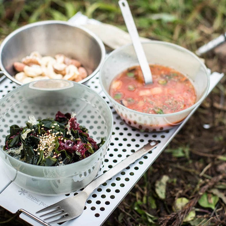 Three bowls sit on a perforated tray outdoors—one filled with leafy greens and sesame seeds, one with soup and a spoon, and one holding beans. A Snow Peak Titanium Fork & Spoon Set rests on the tray, while grass and leaves surround the setup, evoking the practical elegance of an Airstream galley experience.