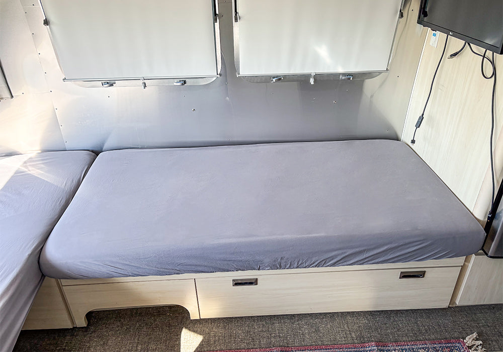 A compact, minimal room features a simple bed dressed with the Airstream Dinette Lounge Cushion Fitted Sheet for Travel Trailers in gray—perfect for custom-fit trailer bedding—set above built-in light wood drawers, with a partial view of a TV mounted on the wall.