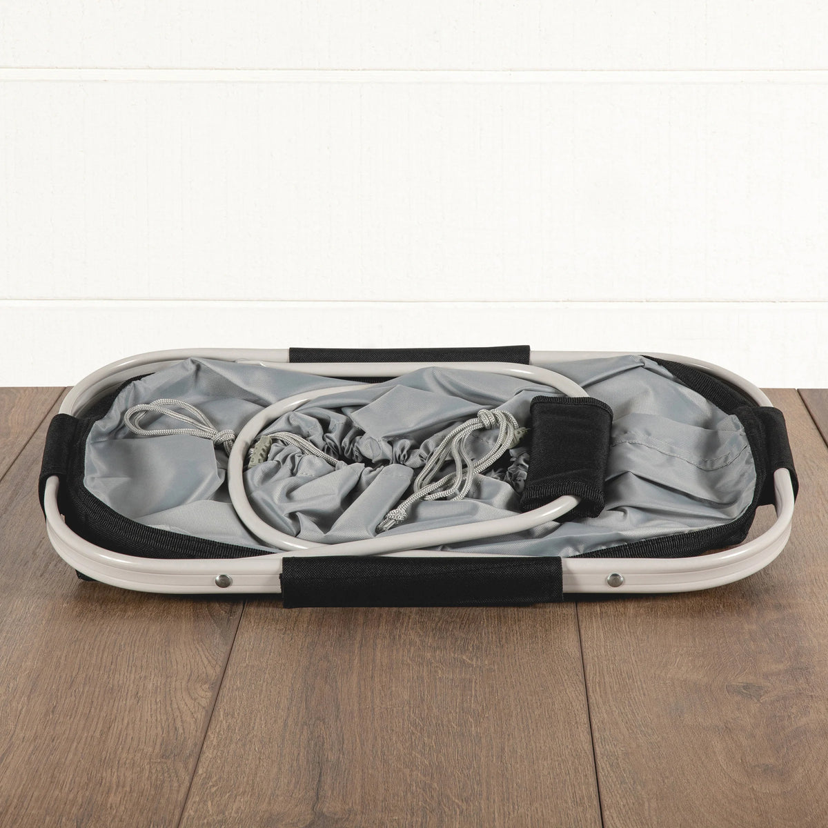 An Airstream Metro Basket Collapsible Cooler Tote in gray, featuring white handles and a collapsible design, lies folded flat on a wooden floor against a white paneled wall.