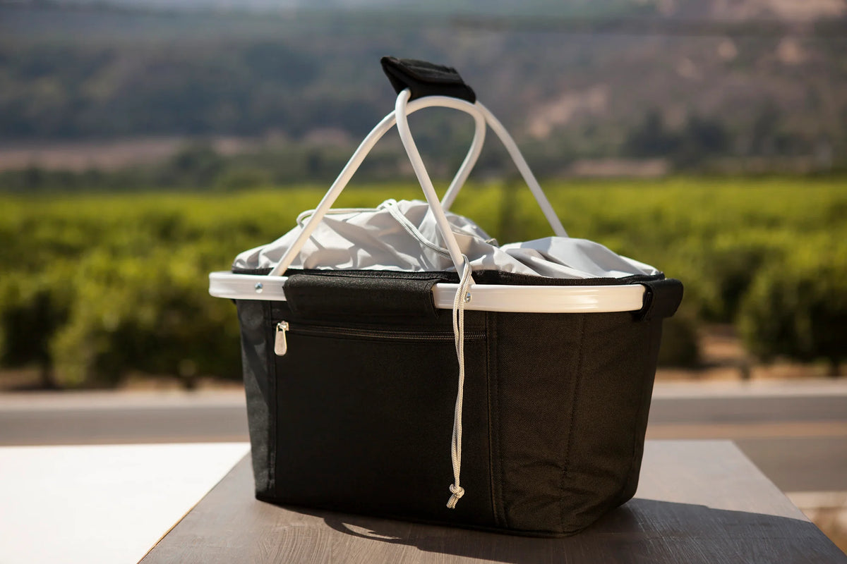An Airstream Metro Basket Collapsible Cooler Tote in a black and white pattern, featuring sturdy handles and a zippered pocket, rests on a wooden outdoor table amid fields and distant hills, serving as both an insulated picnic basket and a convenient collapsible cooler.