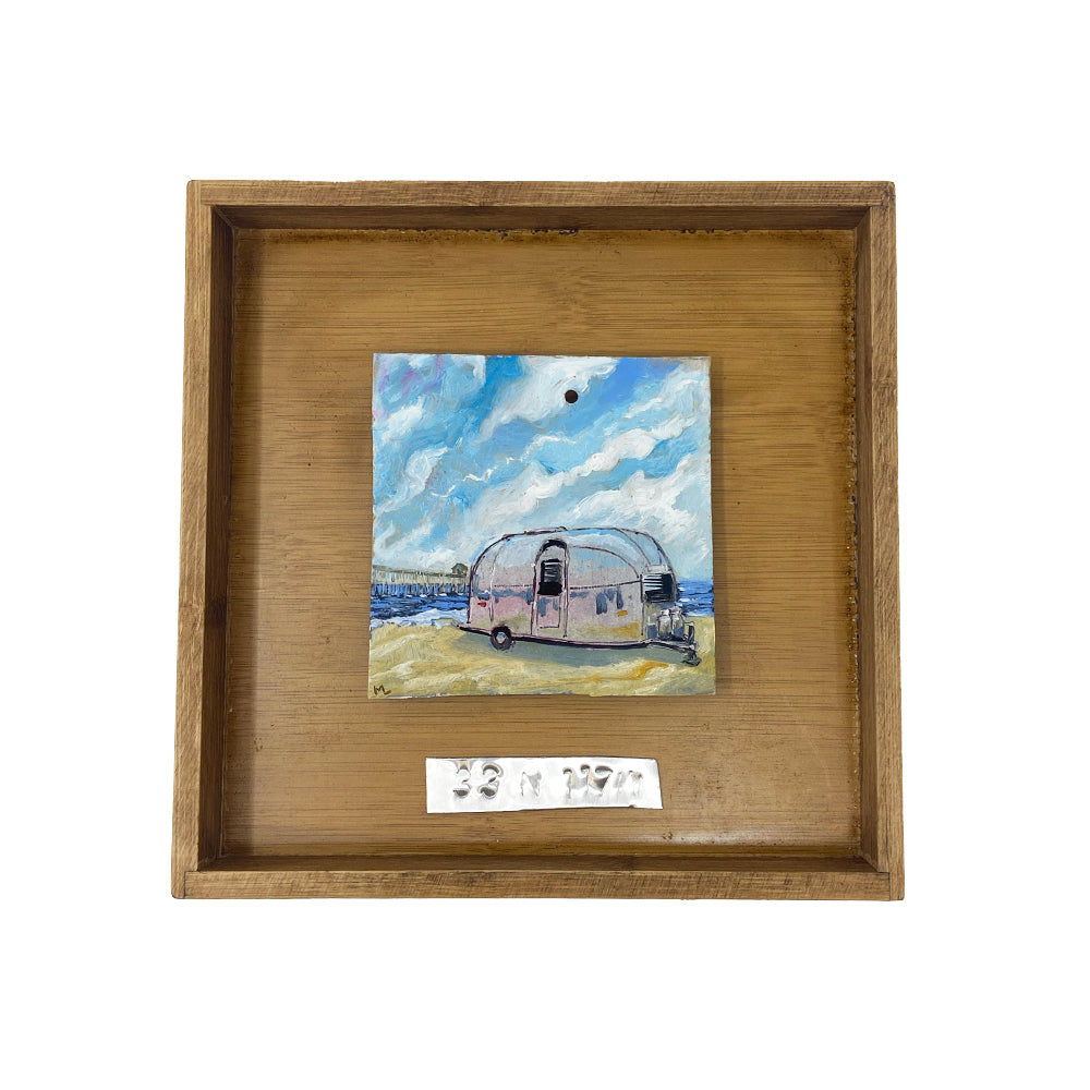 Airstream Original Painting in Shadow Box Frame featuring a silver Airstream trailer on grass beneath a blue sky. The warm wooden shadow box frame beautifully highlights the artwork, and a small metallic label with text is mounted below the painting inside the frame—ideal for those seeking unique salvaged Airstream trailer art and classic Americana wall decor.