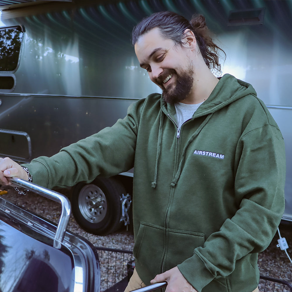 A smiling person in an Airstream Olive Fleece Full Zip Hoodie stands outside near a shiny silver trailer, holding the handle of a grill—an ideal outdoor companion ready for adventure.