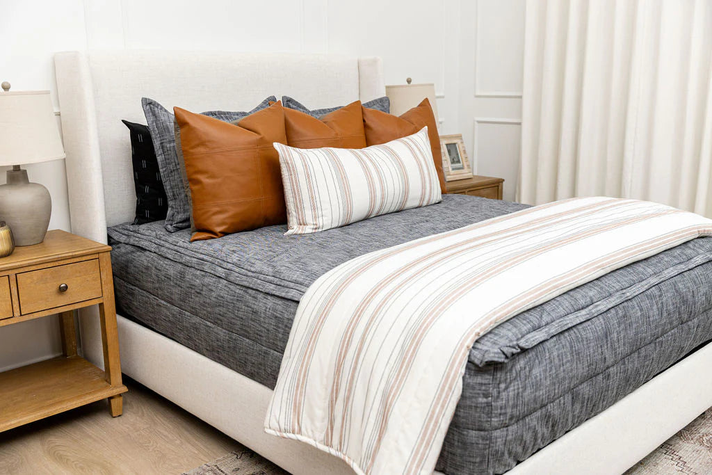 A neatly made bed featuring the Airstream RV Fit Beddy’s for Pottery Barn Travel Trailers bedding, complemented by brown and striped pillows and a coordinating striped blanket. Wooden nightstands with lamps and decor flank the bed in a bright, cozy bedroom.