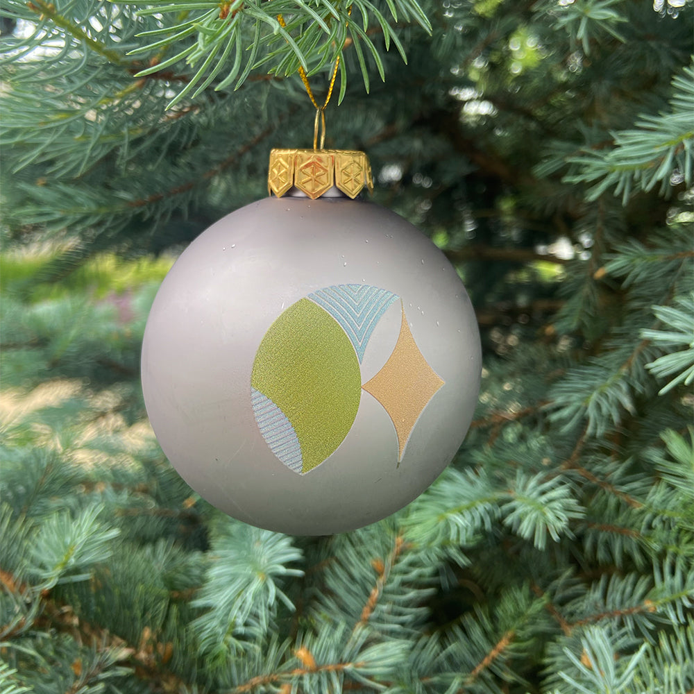 The Frank Lloyd Wright x Airstream Globe Ornament—a silver glass bauble adorned with abstract green, blue, and gold geometric patterns inspired by Frank Lloyd Wright’s iconic style—hangs from a pine tree branch.