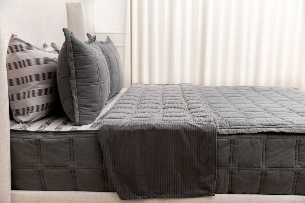 A neatly made bed featuring the Airstream RV Fit Beddy’s for Flying Cloud Travel Trailers—gray quilted bedding with striped and solid pillows and a light-colored upholstered headboard in front of white curtains—highlights the convenience of zippered bedding, ideal for RV mattress or Airstream bedding setups.