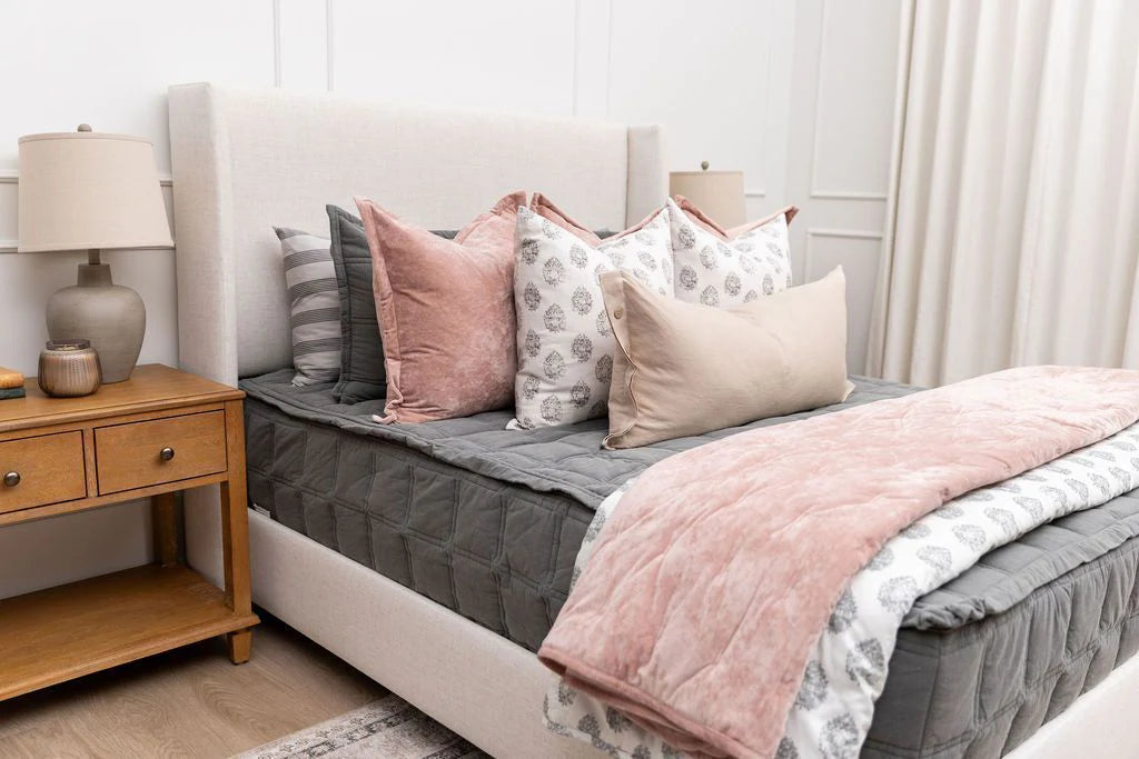 A neatly made bed features the Airstream RV Fit Beddy’s for Flying Cloud Travel Trailers in gray, complemented by a blush pink blanket and multiple decorative pillows. Beside the bed, a wooden nightstand with a lamp and decor completes the look in this bright, modern bedroom.