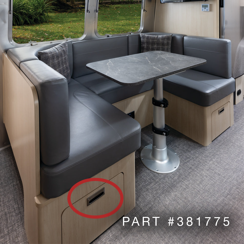 Experience a modern RV dinette featuring dark cushions, a sleek black table, and light wood cabinetry that showcases genuine Airstream craftsmanship. The lower cabinet door is highlighted with a red circle and labeled with “Airstream Interior Flush Door Pull | Part #381775,” emphasizing the premium quality of Airstream cabinet drawers and hardware. Large windows frame vibrant greenery outdoors, completing the inviting atmosphere.