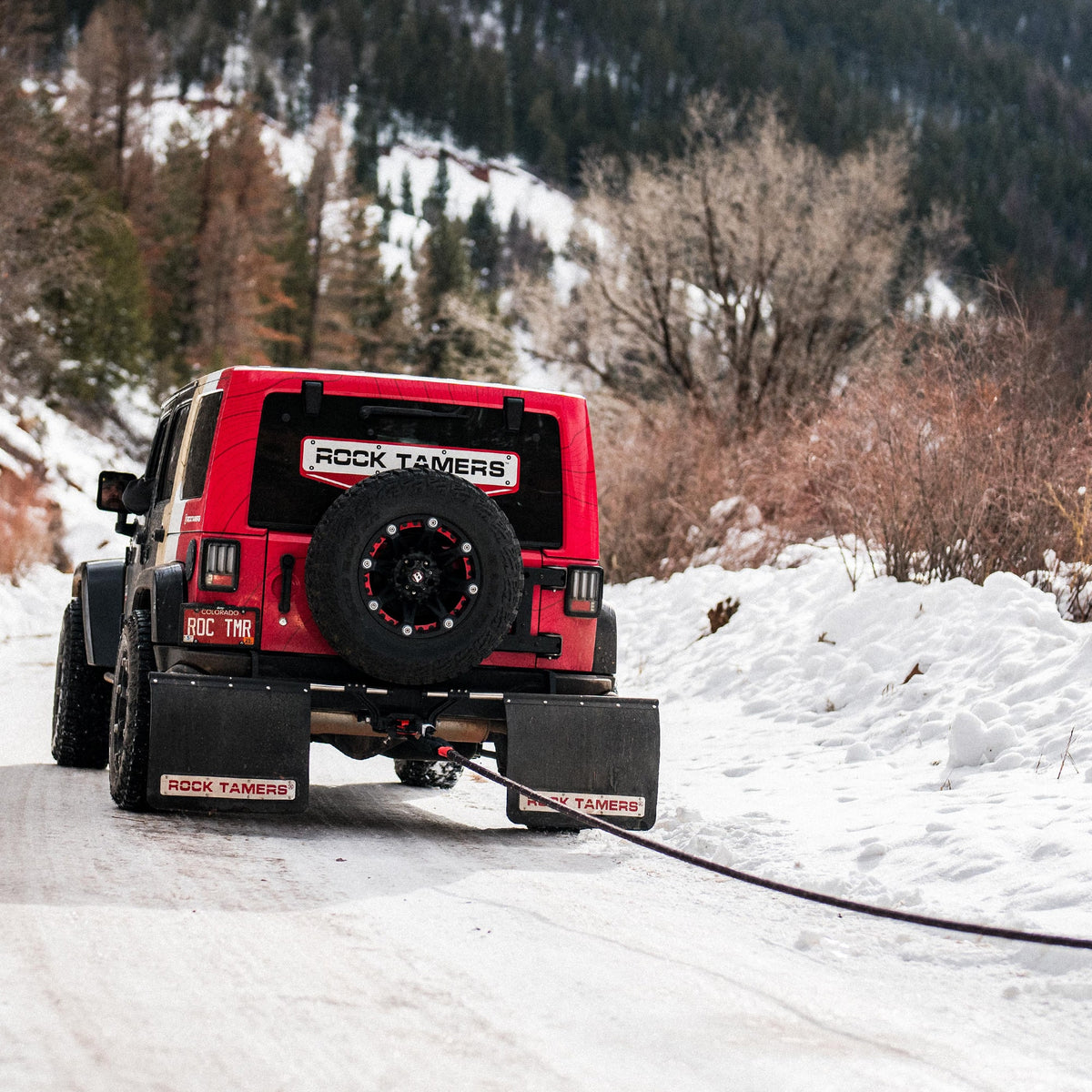 A red Jeep with a spare tire and Rock Tamers mud flaps is parked on a snowy mountain road, facing away. The Rock Tamers Recovery Rope Kit stretches from the back of the vehicle across the snow-covered ground, ready for off-road recovery in challenging winter conditions.