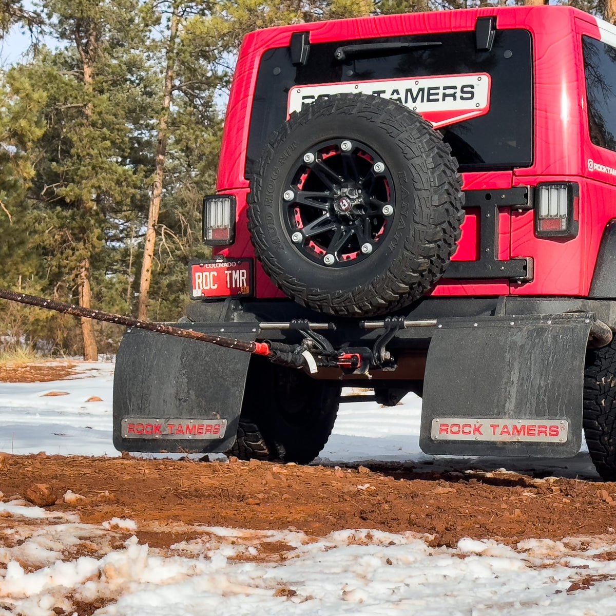 A red off-road vehicle with large mud flaps labeled Rock Tamers is parked on dirt and snow, surrounded by trees. Attached to its rear is the Rock Tamers Recovery Rope Kit, a high-visibility rope indicating it’s being used for hauling or winching during an off-road recovery.